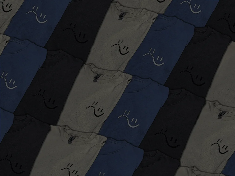 Folded SadImHappy tees in charcoal, olive, and navy, each stamped with our hand-drawn, slightly crooked smiley mark.